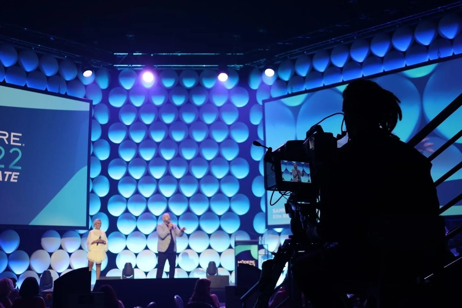 Brett on a lit conference stage, filmed by a camera operator in the foreground